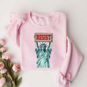 Op de afbeelding: Roze sweatshirt met een afbeelding van het Vrijheidsbeeld dat een bord vasthoudt met de tekst "RESIST".