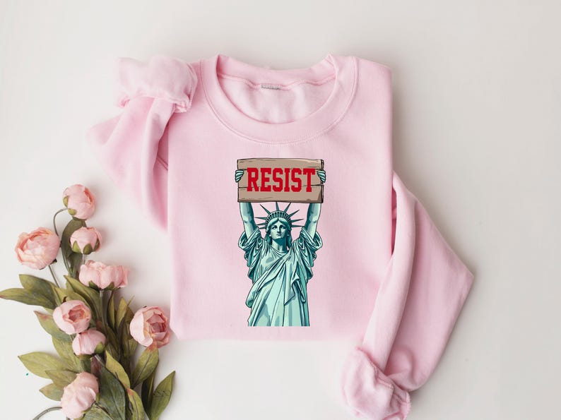 Op de afbeelding: Roze sweatshirt met een afbeelding van het Vrijheidsbeeld dat een bord vasthoudt met de tekst "RESIST".