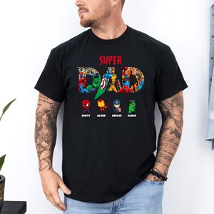 May include: Black t-shirt featuring the text "SUPER DAD" in red, with a collage of superhero characters forming the letters. Below, cartoon superheroes are named: Amity, Alger, Devlin, and Alvar. A casual tee for fans.