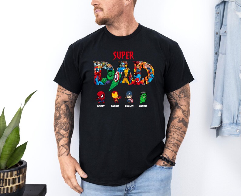 May include: Black t-shirt featuring the text "SUPER DAD" in red, with a collage of superhero characters forming the letters. Below, cartoon superheroes are named: Amity, Alger, Devlin, and Alvar. A casual tee for fans.