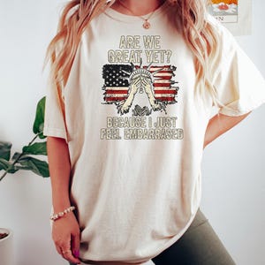 Puede incluir: Una camiseta blanca con un gr&aacute;fico de bandera estadounidense desgastado y el texto "Are we great yet? Because I just feel embarrassed."