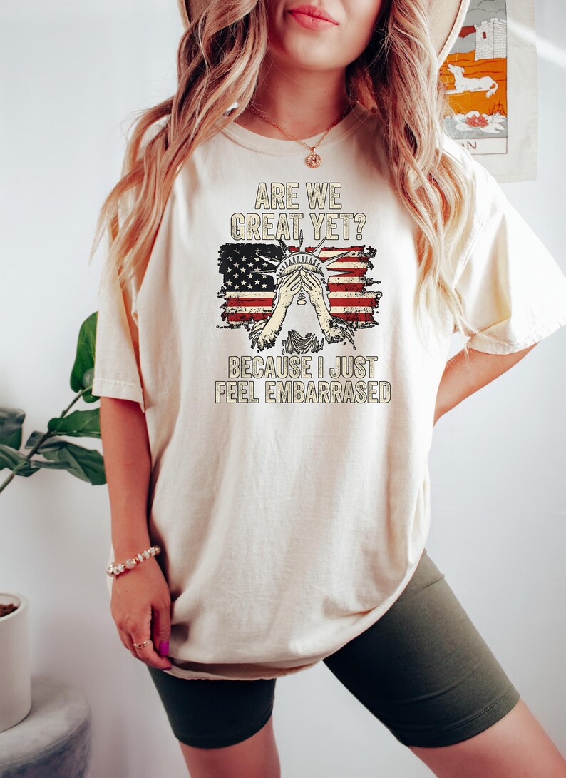 Puede incluir: Una camiseta blanca con un gr&aacute;fico de bandera estadounidense desgastado y el texto "Are we great yet? Because I just feel embarrassed."