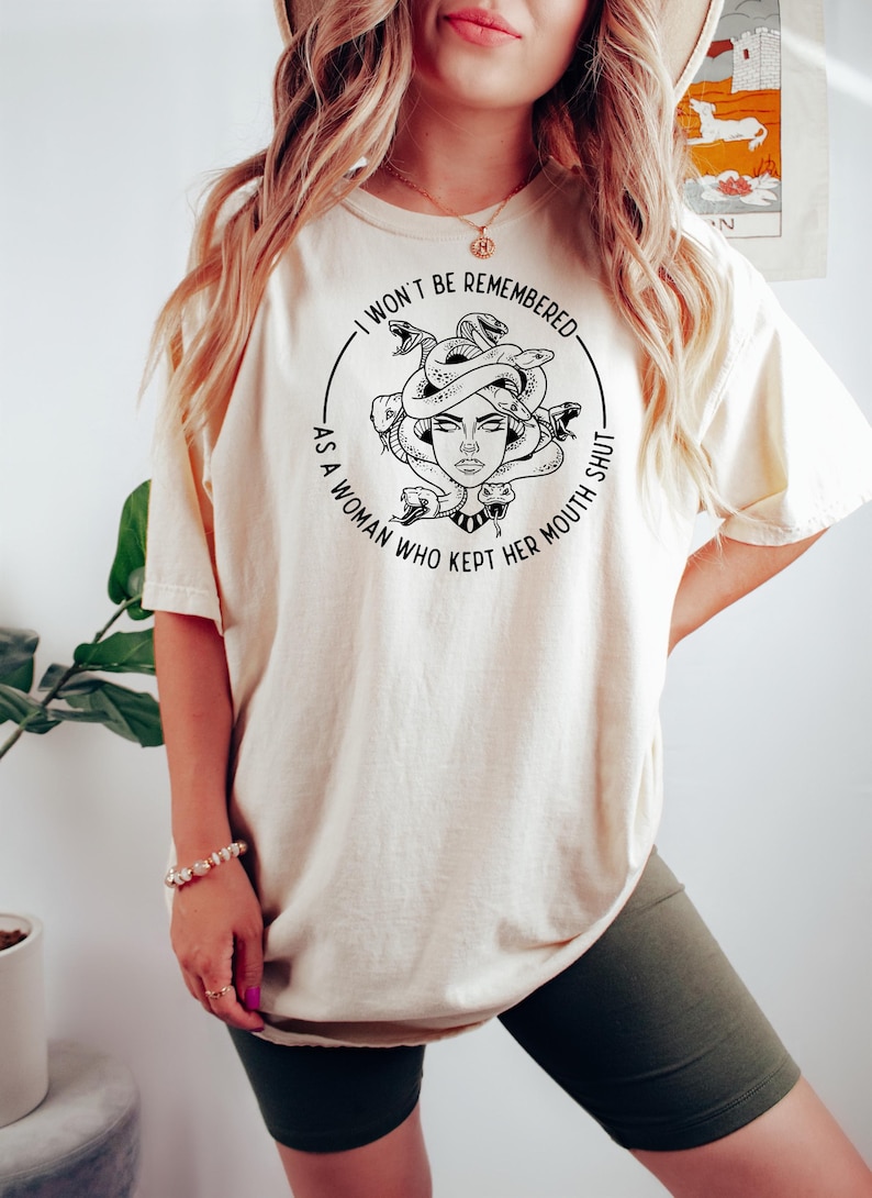 May include: Cream-colored t-shirt featuring a black and white illustration of Medusa with snakes for hair. The text around the image reads, "I WON'T BE REMEMBERED AS A WOMAN WHO KEPT HER MOUTH SHUT."