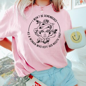 May include: A light pink sweatshirt with a Medusa graphic and the text "I WON'T BE REMEMBERED AS A WOMAN WHO KEPT HER MOUTH SHUT." The sweatshirt is paired with light blue shorts. A white mug with a smiley face is held in the hand.