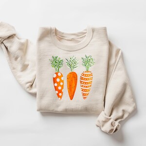 Puede incluir: Una sudadera beige con un gr&aacute;fico de tres zanahorias naranjas con lunares blancos y rayas. Las zanahorias tienen hojas verdes.