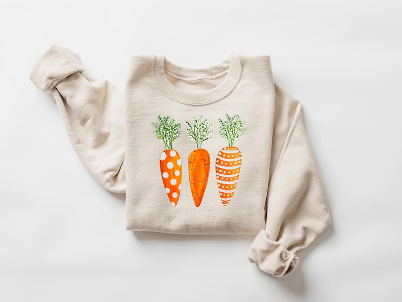 Puede incluir: Una sudadera beige con un gr&aacute;fico de tres zanahorias naranjas con lunares blancos y rayas. Las zanahorias tienen hojas verdes.