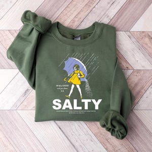May include: An olive green sweatshirt with a graphic of a person under a blue umbrella, with the word "SALTY" in white. The person is wearing a yellow dress and is sprinkling salt. The text "All those flavors and you choose to be" is also present.