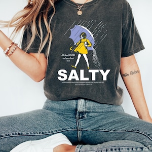 May include: Dark gray t-shirt with the word "SALTY" in large white letters. The shirt features a graphic of a person under an umbrella, with text that reads "All the flavors and you choose to be."