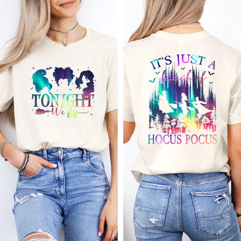 May include: Cream-colored t-shirt featuring a colorful graphic with the text "TONIGHT We fly" and silhouettes of three witches. The back of the shirt displays the text "IT'S JUST A bunch of HOCUS POCUS" with witch silhouettes.