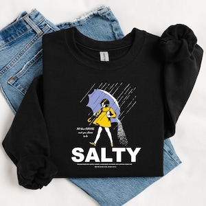 May include: Black sweatshirt with a graphic of a person under a blue umbrella, with the word "SALTY" in large white letters. The person is wearing a yellow coat and the image includes the text "All the flavors and you choose to be".