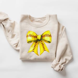 K&ouml;nnte beinhalten: Ein beiger Pullover mit einem gelben und roten Softball-Schleifendesign.