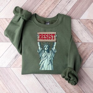 Op de afbeelding: Groen sweatshirt met een afbeelding van het Vrijheidsbeeld dat een bord vasthoudt met de tekst "RESIST".