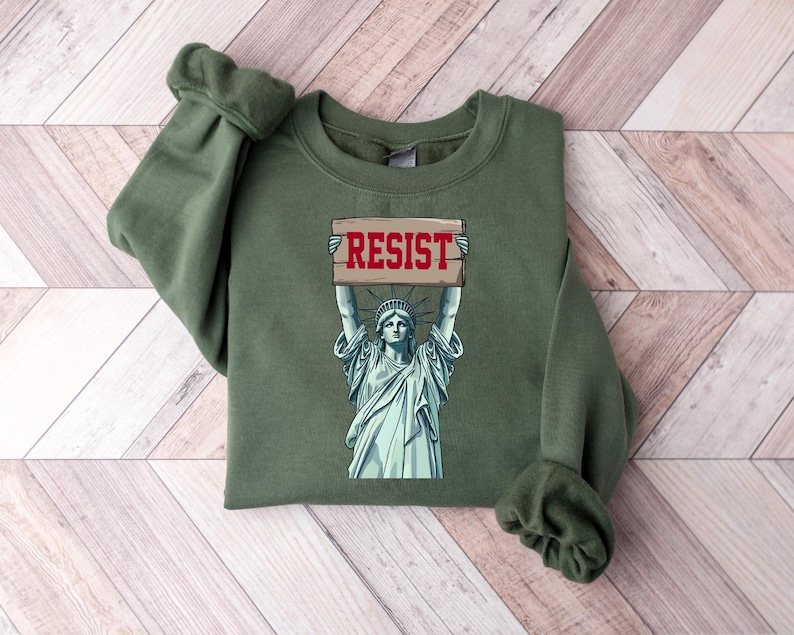 Op de afbeelding: Groen sweatshirt met een afbeelding van het Vrijheidsbeeld dat een bord vasthoudt met de tekst "RESIST".