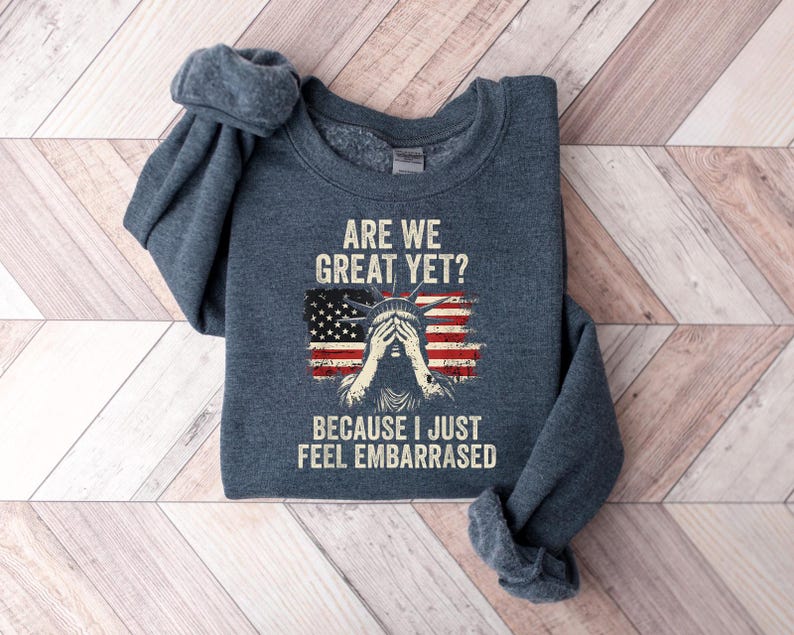 Puede incluir: Una sudadera gris oscura con el texto "Are we great yet? Because I just feel embarrassed" y un gr&aacute;fico de la Estatua de la Libertad con la bandera estadounidense detr&aacute;s de ella.