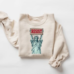Op de afbeelding: Een beige sweatshirt met een afbeelding van het Vrijheidsbeeld dat een bord vasthoudt met de tekst "RESIST".