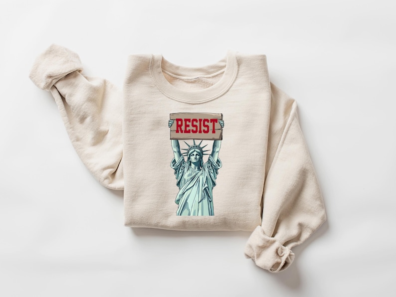 Op de afbeelding: Een beige sweatshirt met een afbeelding van het Vrijheidsbeeld dat een bord vasthoudt met de tekst "RESIST".