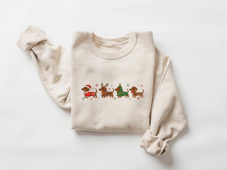 May include: Beige crewneck sweatshirt with a festive dachshund design. The graphic features four dachshunds dressed in holiday attire, including a Santa hat, reindeer antlers, and a Christmas tree costume. The design is accented with snowflakes.