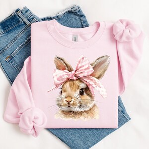 May include: Pink sweatshirt with a brown bunny wearing a pink and white bow. The bunny is looking to the right.