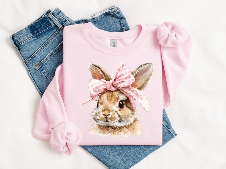 May include: Pink sweatshirt with a brown bunny wearing a pink and white bow. The bunny is looking to the right.
