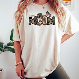 May include: A white t-shirt with the text "army mom" in camouflage print with a pair of army boots and a small American flag.