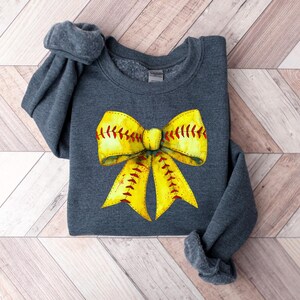K&ouml;nnte beinhalten: Ein dunkelgraues Sweatshirt mit einem gelben und roten Softball-Schleifendesign. Die Schleife ist so gestaltet, dass sie einem Softball mit roten N&auml;hten &auml;hnelt.