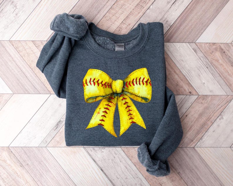 K&ouml;nnte beinhalten: Ein dunkelgraues Sweatshirt mit einem gelben und roten Softball-Schleifendesign. Die Schleife ist so gestaltet, dass sie einem Softball mit roten N&auml;hten &auml;hnelt.