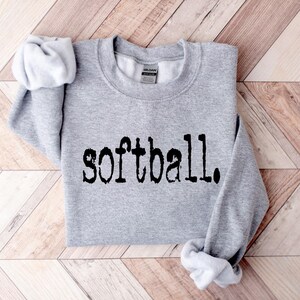 K&ouml;nnte beinhalten: Ein grauer Pullover mit dem Wort "Softball" in Schwarz auf der Vorderseite.