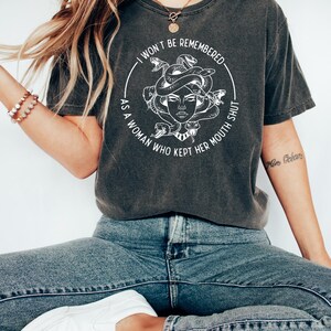 May include: Dark gray t-shirt featuring a white graphic of Medusa with snakes and the text "I WON'T BE REMEMBERED AS A WOMAN WHO KEPT HER MOUTH SHUT." The shirt is paired with blue jeans and white sneakers.