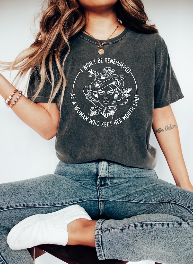 May include: Dark gray t-shirt featuring a white graphic of Medusa with snakes and the text "I WON'T BE REMEMBERED AS A WOMAN WHO KEPT HER MOUTH SHUT." The shirt is paired with blue jeans and white sneakers.