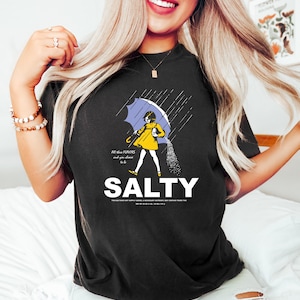 May include: Black t-shirt with a graphic of a person under a blue umbrella, with salt pouring down. The word "SALTY" is printed in large white letters below the graphic. Text on the shirt reads "All these flavors and you choose to be."