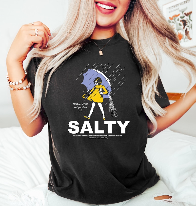 May include: Black t-shirt with a graphic of a person under a blue umbrella, with salt pouring down. The word "SALTY" is printed in large white letters below the graphic. Text on the shirt reads "All these flavors and you choose to be."