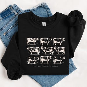 Op de afbeelding: Zwart sweatshirt met een raster van negen witte koeienillustraties. De koeien hebben verschillende zwart-witte patronen. De tekst "SUPPORT YOUR LOCAL FARMER" staat eronder gedrukt. Het sweatshirt wordt getoond op een blauwe spijkerbroek.