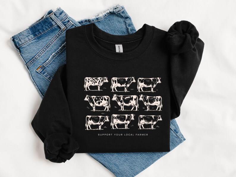 Op de afbeelding: Zwart sweatshirt met een raster van negen witte koeienillustraties. De koeien hebben verschillende zwart-witte patronen. De tekst "SUPPORT YOUR LOCAL FARMER" staat eronder gedrukt. Het sweatshirt wordt getoond op een blauwe spijkerbroek.