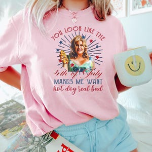 May include: A pink t-shirt with a graphic of a woman holding a hot dog and the text "You look like the 4th of July makes me want hot dog real bad".