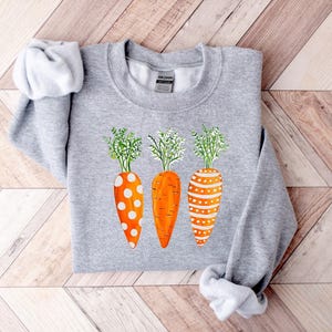 Puede incluir: Una sudadera gris con un dise&ntilde;o de tres zanahorias naranjas con lunares blancos y rayas. Las zanahorias tienen hojas verdes.