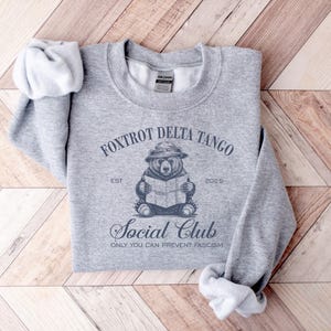 Puede incluir: Sudadera gris con un gr&aacute;fico de un oso con un sombrero leyendo un libro. El texto "Foxtrot Delta Tango Est 2025 Social Club Only You Can Prevent Fascism" est&aacute; impreso en la sudadera.