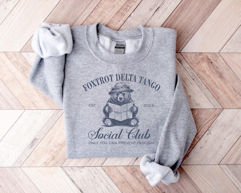 Puede incluir: Sudadera gris con un gr&aacute;fico de un oso con un sombrero leyendo un libro. El texto "Foxtrot Delta Tango Est 2025 Social Club Only You Can Prevent Fascism" est&aacute; impreso en la sudadera.