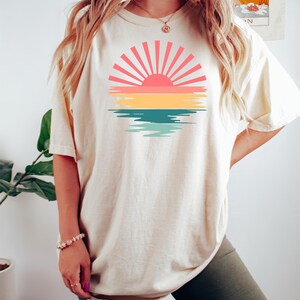 May include: A white oversized t-shirt with a retro sunset graphic featuring a pink sun with yellow, green, and blue water reflections. The model is wearing the shirt with olive green biker shorts.