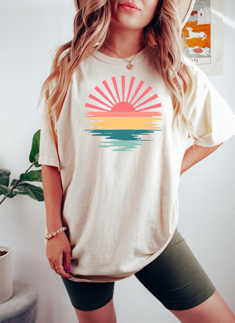 May include: A white oversized t-shirt with a retro sunset graphic featuring a pink sun with yellow, green, and blue water reflections. The model is wearing the shirt with olive green biker shorts.