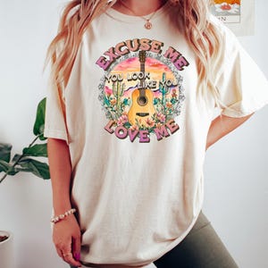 May include: A cream-colored oversized t-shirt with a graphic print of a sunset, cactus, guitar, and flowers. The text on the shirt reads "Excuse Me You Look Like You Love Me".