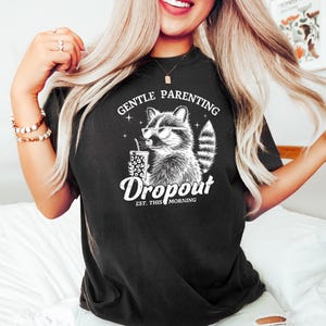 May include: Black t-shirt featuring a white graphic of a raccoon wearing sunglasses and holding a cup. The text "Gentle Parenting Dropout Est. This Morning" is printed below the raccoon.