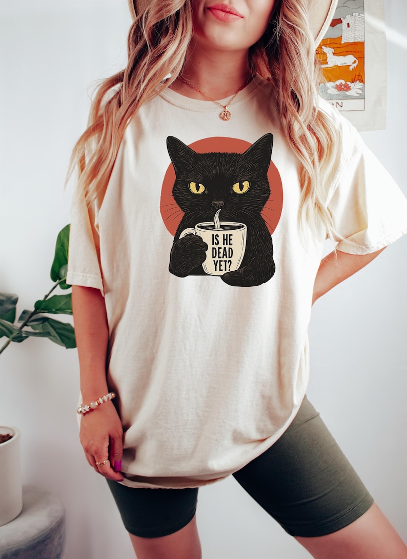 May include: A white t-shirt with a black cat illustration holding a coffee mug that says "Is he dead yet?" The cat is looking at the mug with a serious expression.