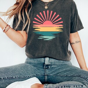 May include: A dark gray t-shirt with a vintage-style sunset graphic featuring a red, orange, yellow, green, and blue color scheme. The graphic depicts a sun with rays and a reflection in the water.