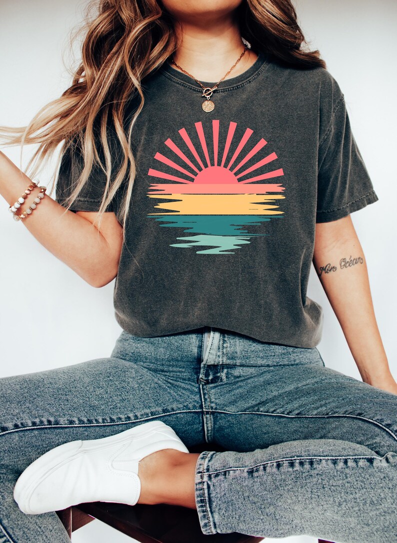 May include: A dark gray t-shirt with a vintage-style sunset graphic featuring a red, orange, yellow, green, and blue color scheme. The graphic depicts a sun with rays and a reflection in the water.
