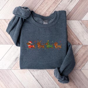 May include: A dark gray sweatshirt featuring an embroidered design of four dachshunds dressed in Christmas attire. The dachshunds are wearing a Santa hat, reindeer antlers, a Christmas tree costume, and a festive scarf. The design is in shades of red, green, and brown.
