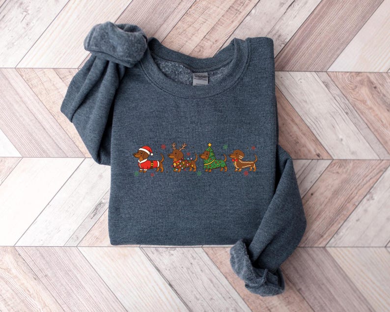 May include: A dark gray sweatshirt featuring an embroidered design of four dachshunds dressed in Christmas attire. The dachshunds are wearing a Santa hat, reindeer antlers, a Christmas tree costume, and a festive scarf. The design is in shades of red, green, and brown.