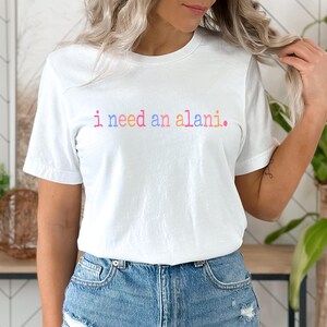 May include: A white t-shirt with the text "i need an alani." in rainbow colors.
