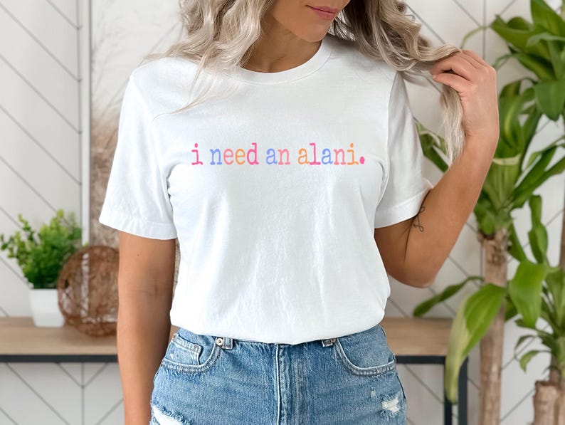 May include: A white t-shirt with the text "i need an alani." in rainbow colors.