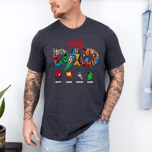 May include: Dark grey t-shirt featuring the text "SUPER DAD" in a colourful comic book design. Below the text are cartoon superhero images with names: Amity, Alder, Devlin, and Alvar. The shirt has a crew neck.