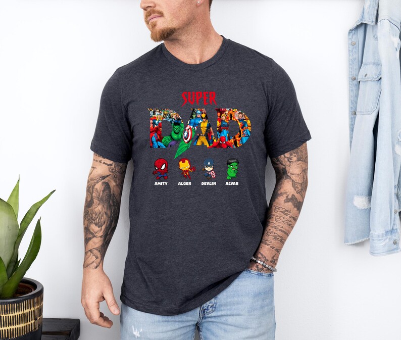 May include: Dark grey t-shirt featuring the text "SUPER DAD" in a colourful comic book design. Below the text are cartoon superhero images with names: Amity, Alder, Devlin, and Alvar. The shirt has a crew neck.
